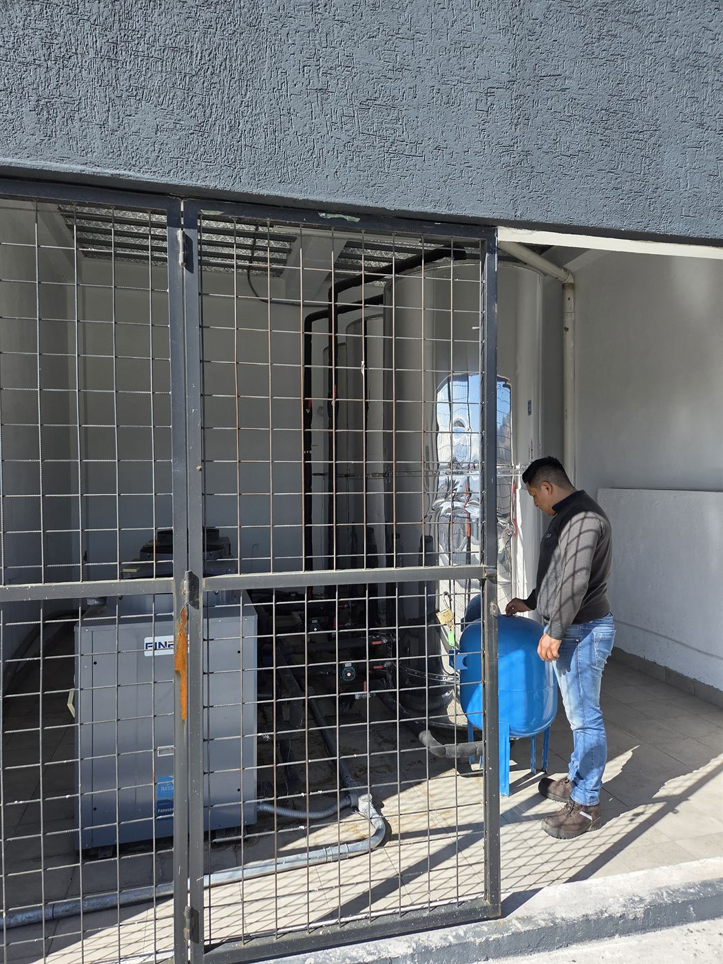 Técnico junto a un acumulador de agua caliente y un tanque hidroneumático azul en un cuarto de máquinas